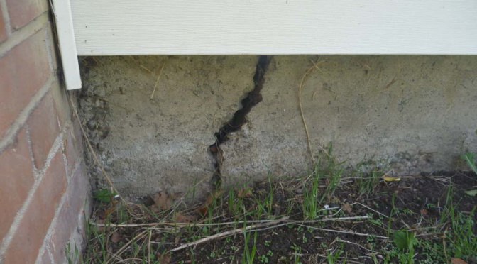 This is an image that shows a section of brick wall beside a slab of concrete foundation with a large thick crack in it that needs to be addressed by professionals.